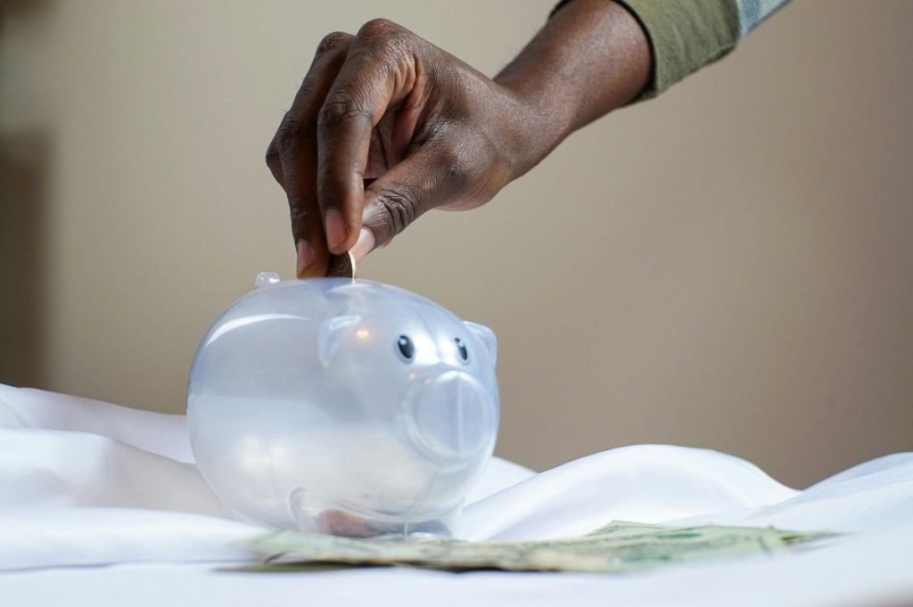 Mão depositando moeda em cofrinho transparente para Colchão de Segurança (Reserva de Emergência).
